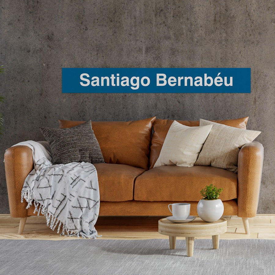 Sofá marrón con cojines, manta y letrero azul Santiago Bernabéu en pared gris