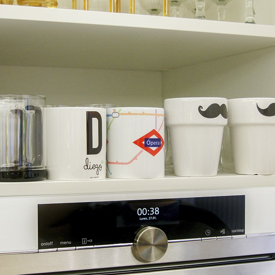 Tazas originales en estante de cocina, una con mapa metro Opera y otras con bigote, junto a horno