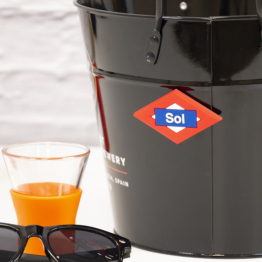 Cubo negro Sol, vaso naranja y gafas de sol sobre mesa blanca, fondo desenfocado