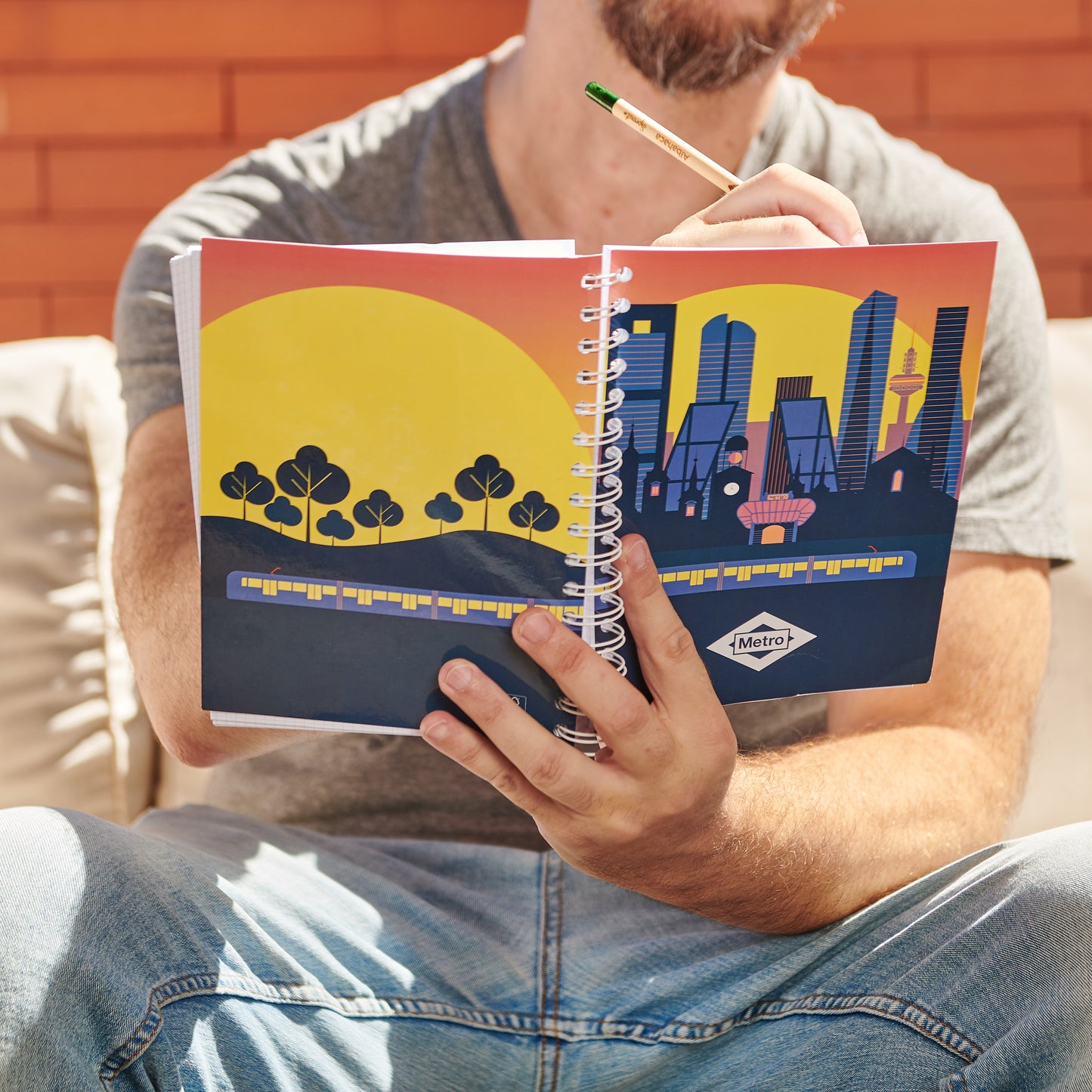 Hombre sostiene cuaderno Metro con ilustración de ciudad al atardecer y tren amarillo