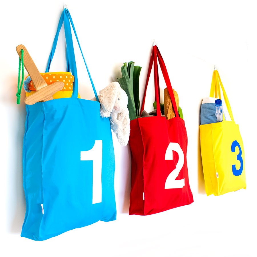 Bolsas reutilizables de tela azul, roja y amarilla numeradas, colgadas en pared blanca.