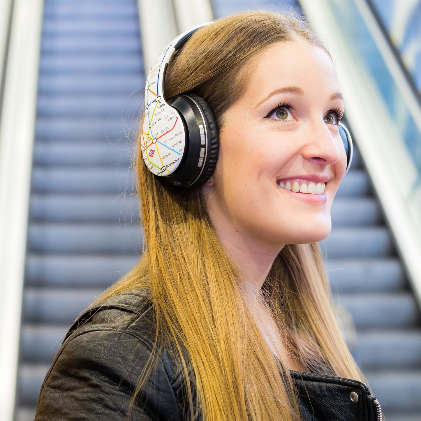 Mujer joven con auriculares decorados con mapa, sonriendo en una escalera eléctrica.