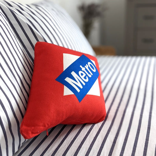 Cojín rojo con logo Metro sobre cama con sábanas blancas de rayas negras