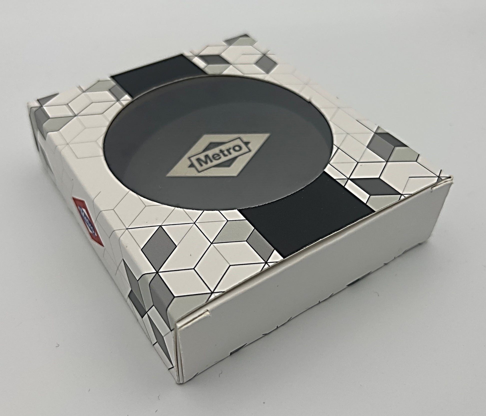 Caja decorativa blanca con patrón geométrico, ventana circular y logo Metro al centro