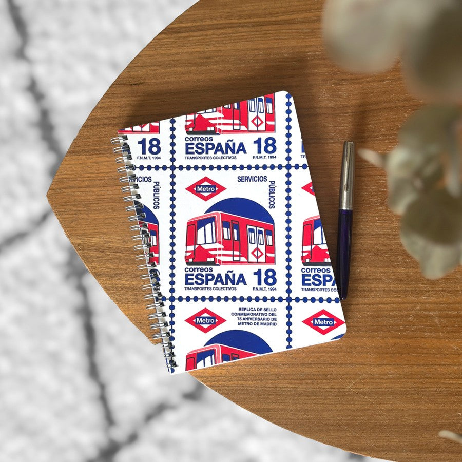 Libreta con diseño de sello de metro de Madrid sobre mesa de madera junto a bolígrafo.