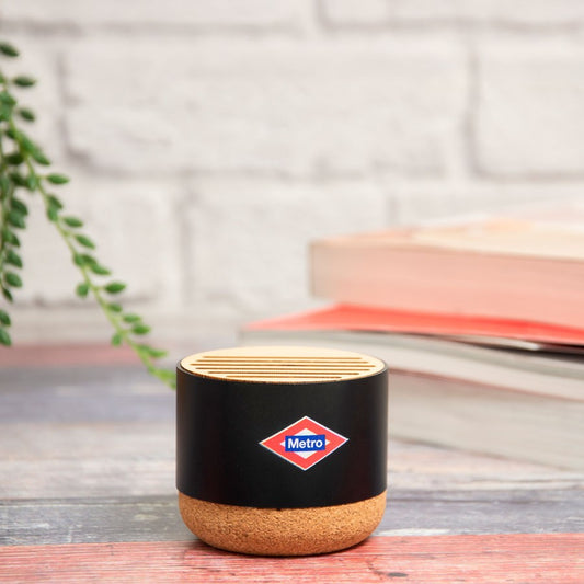 Altavoz Bluetooth negro Metro sobre mesa de madera, libros y planta al fondo, decoración moderna.