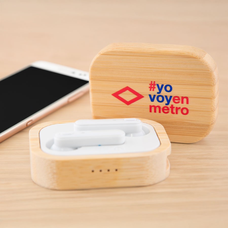 Audífonos inalámbricos blancos en estuche de bambú con hashtag #yo voy en metro, junto a smartphone.