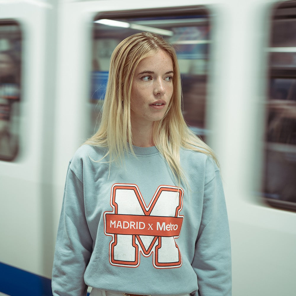 Chica rubia con sudadera azul de Madrid Metro en estación de metro con tren en movimiento