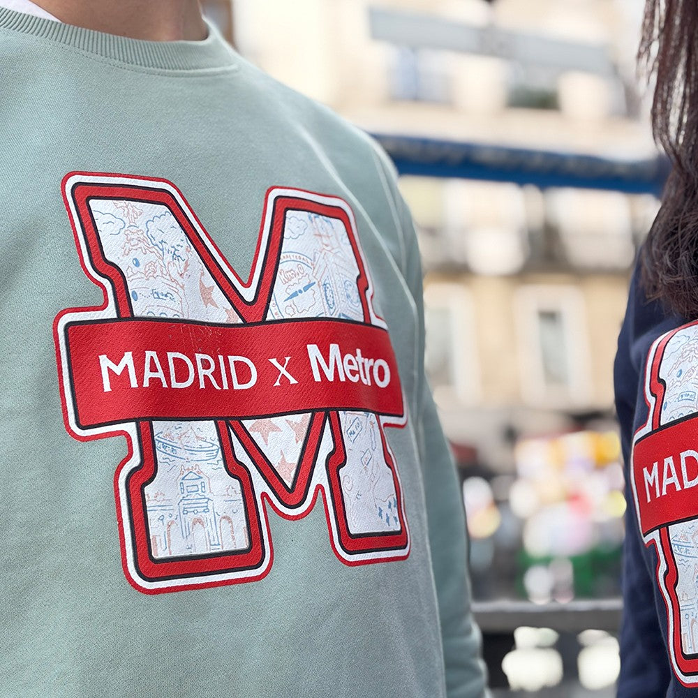 Sudadera verde con letra M y logo Madrid x Metro, diseño urbano, personas en exterior urbano.