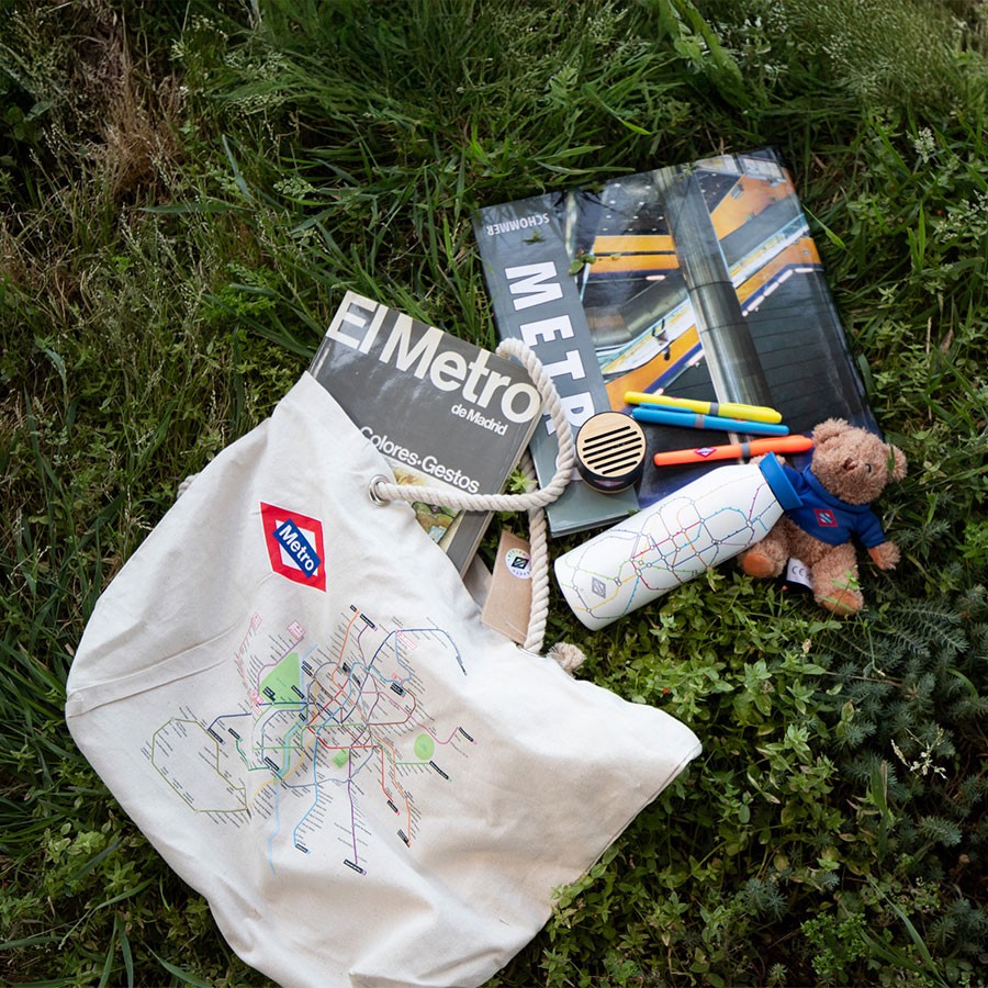 Bolsa, botella y libros del Metro de Madrid sobre césped, junto a bolígrafos y peluche.