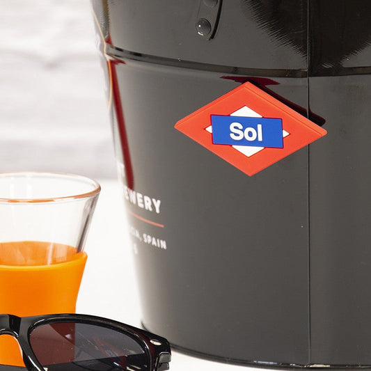 Cubo negro Sol, vaso naranja y gafas de sol sobre mesa blanca, fondo desenfocado