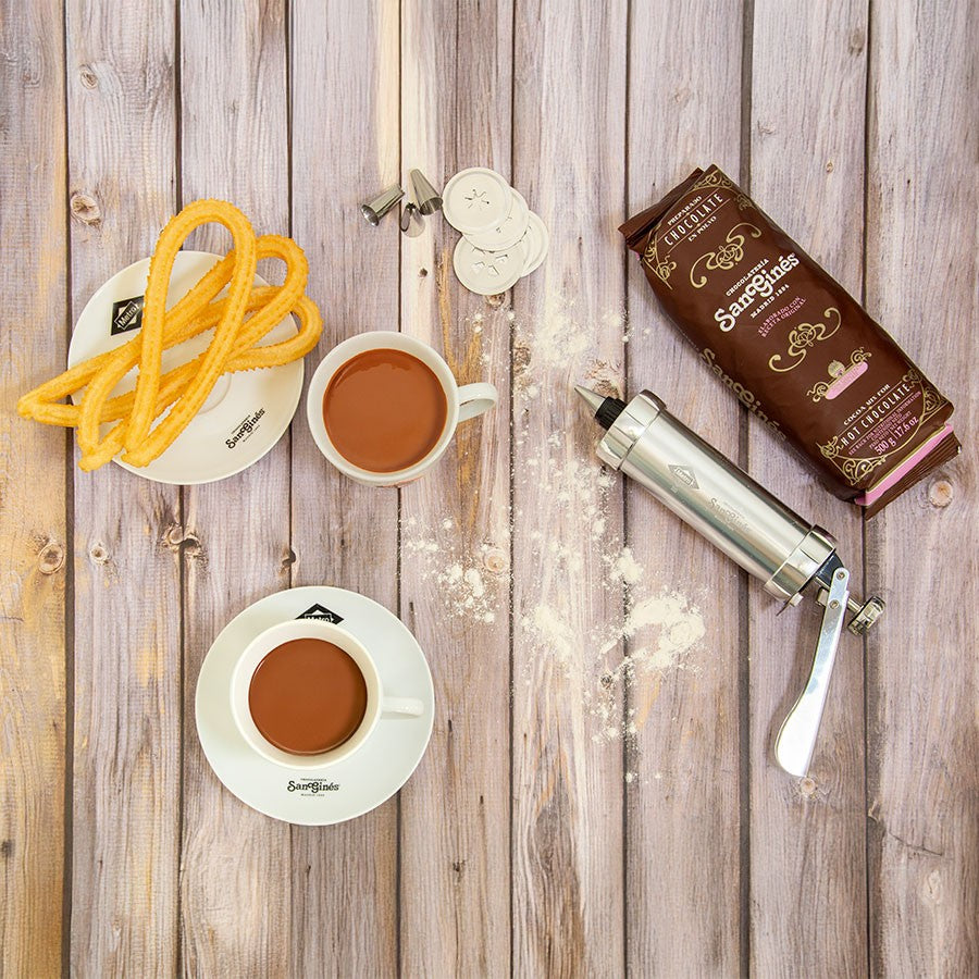 Chocolate caliente con churros y churrera sobre mesa de madera, paquete de chocolate Sanguínes.