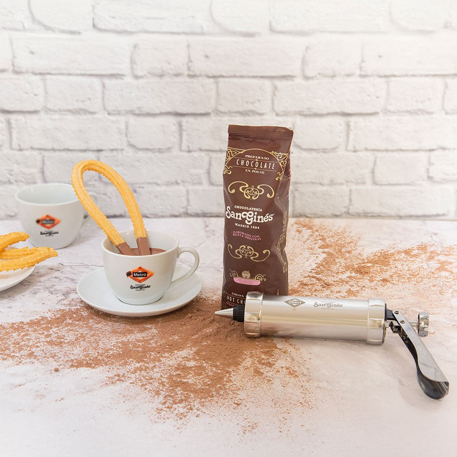 Churros en taza de chocolate San Ginés, manga pastelera y cacao sobre mesa blanca