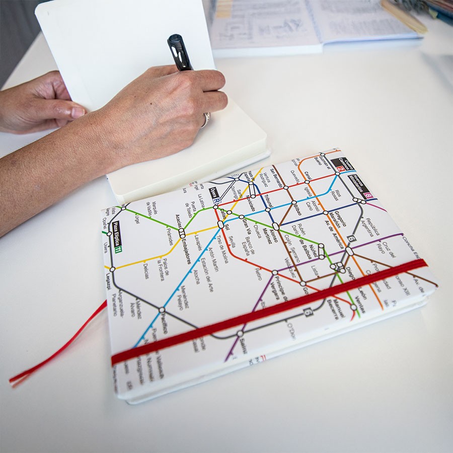 Persona escribiendo en libreta junto a cuaderno con diseño de mapa metro Madrid sobre mesa blanca