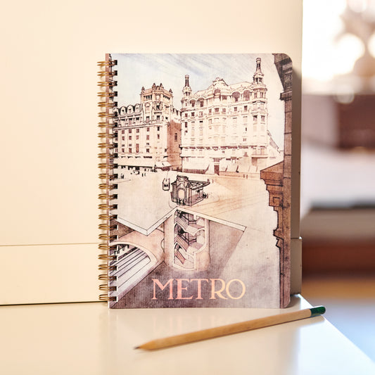 Cuaderno con portada de dibujo arquitectónico del metro y edificios históricos, sobre mesa blanca con lápiz.