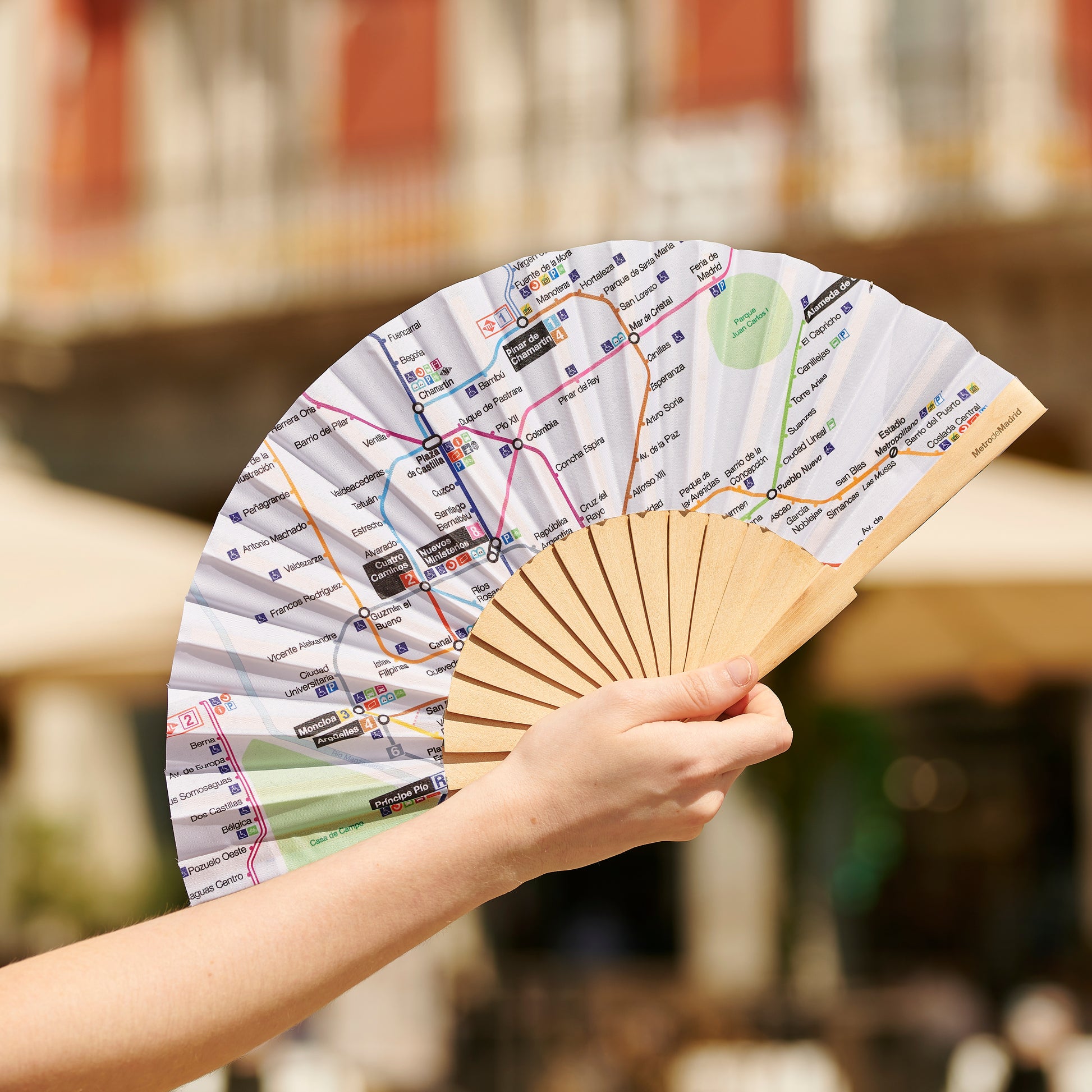 Abanico de papel con mapa del metro de Madrid sostenido por una mano en exterior urbano.