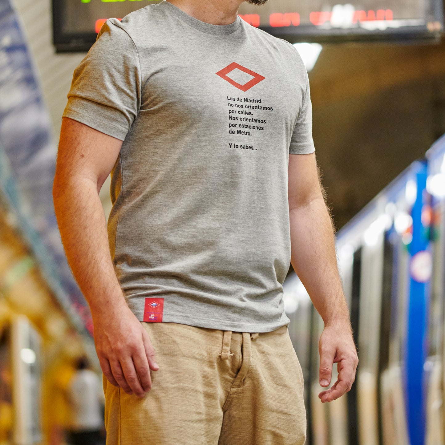 Hombre con camiseta gris con texto de Madrid en estación de metro, tren azul de fondo