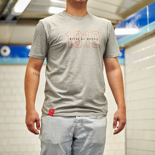 Camiseta gris Metro de Madrid 1919, hombre en estación de metro moderna.