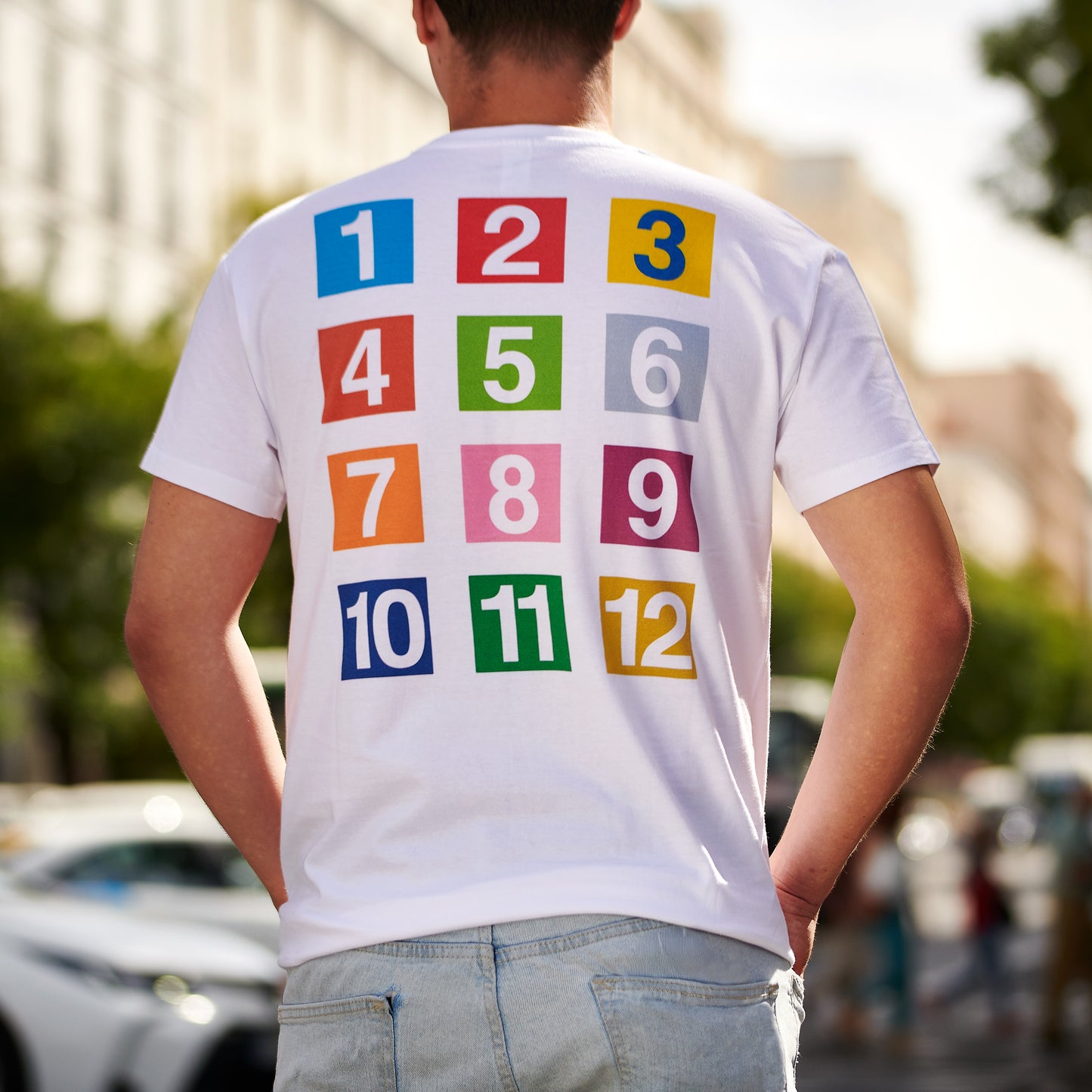 Hombre con camiseta blanca de números coloridos del 1 al 12, fondo urbano y soleado