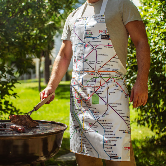 Hombre asando carne con delantal estampado del mapa del metro de Madrid en jardín veraniego
