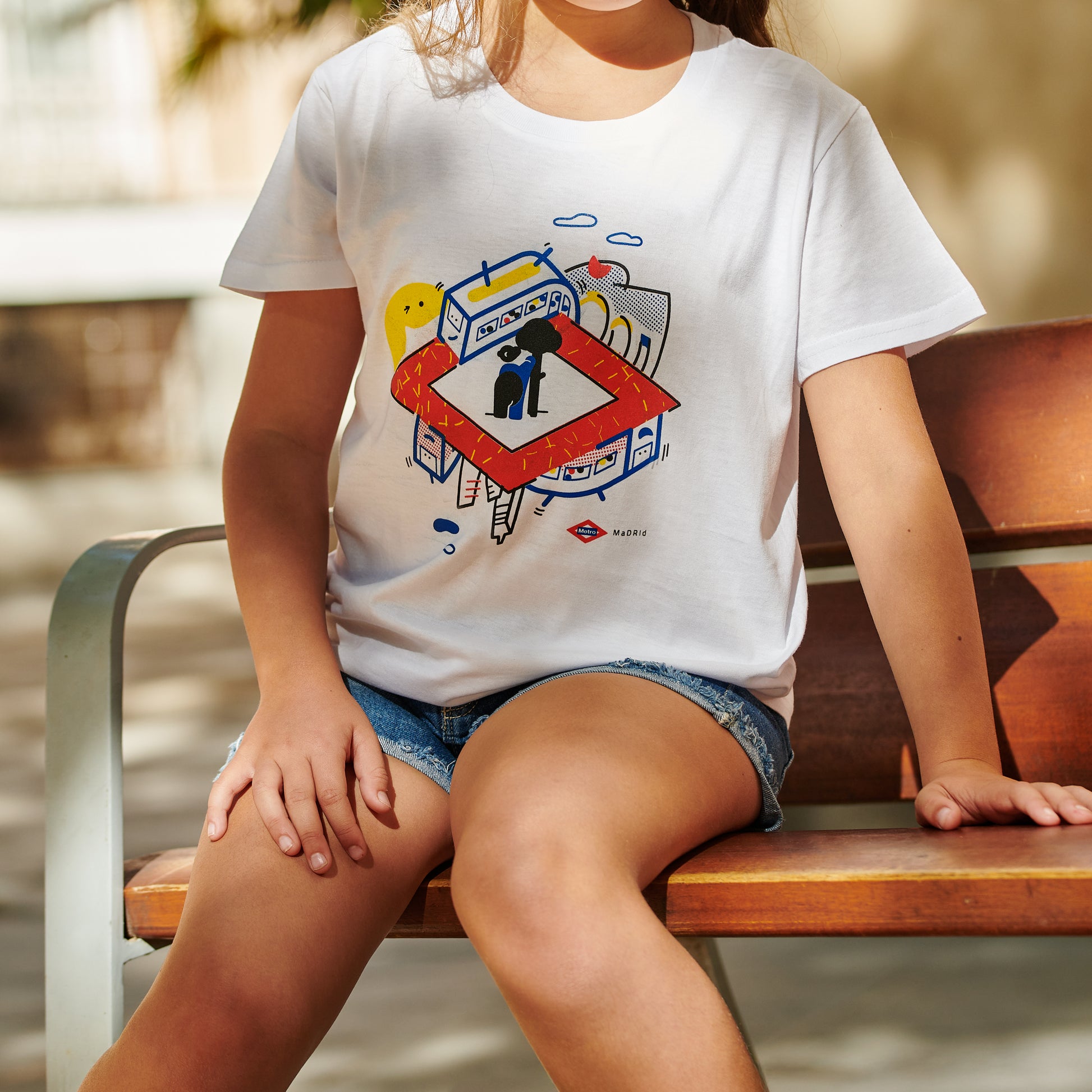 Niña sentada en banco con camiseta blanca de diseño Madrid, ilustración colorida y shorts de mezclilla